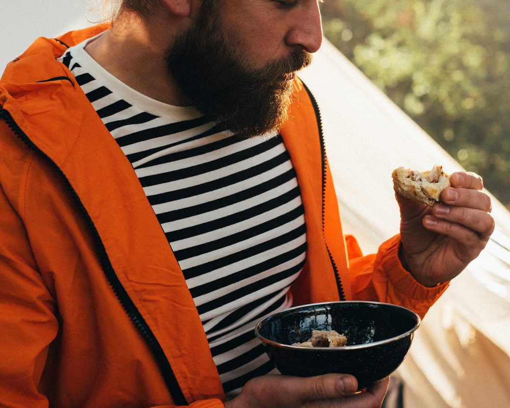Man in an orange jacket eating a pork pie with a bowl of chutney outdoors
