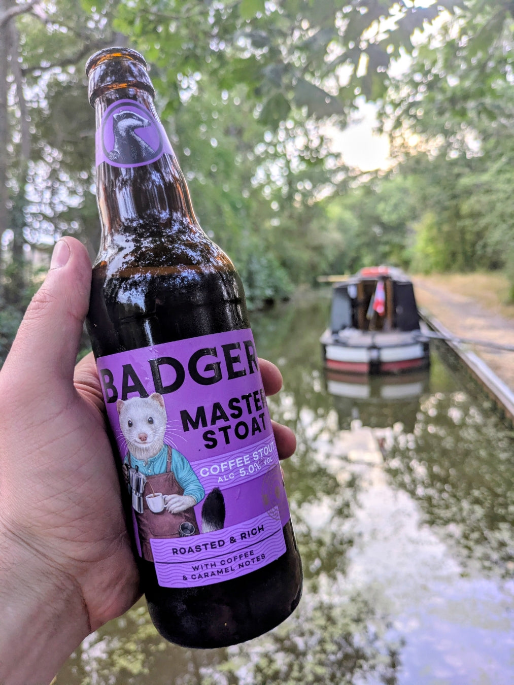 Hand holding a bottle of Badger Master Stoat beside a canal with narrowboats