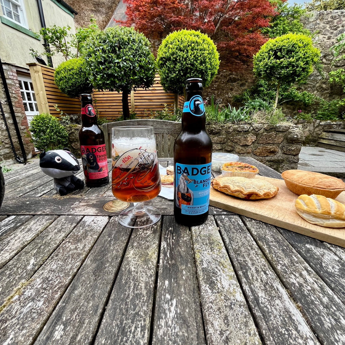 Two bottles of Badger beers (Tangle Foot and Blandford Fly) and a filled pint glass on a weathered wooden table in a garden setting, with pastries on a cutting board and a small badger figurine nearby.