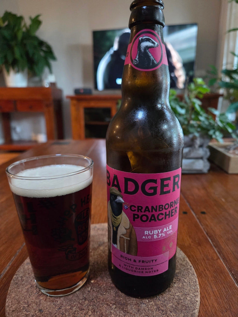 A bottle of Badger Cranborne Poacher Ruby Ale next to a filled pint glass on a coaster, set on a wooden table in a living room background, with plants in background.