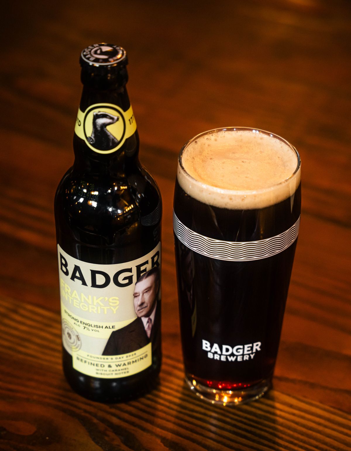 Bottle of Badger Frank's Integrity alongside poured pint of dark ale in Badger pint glass, against dark wooden table