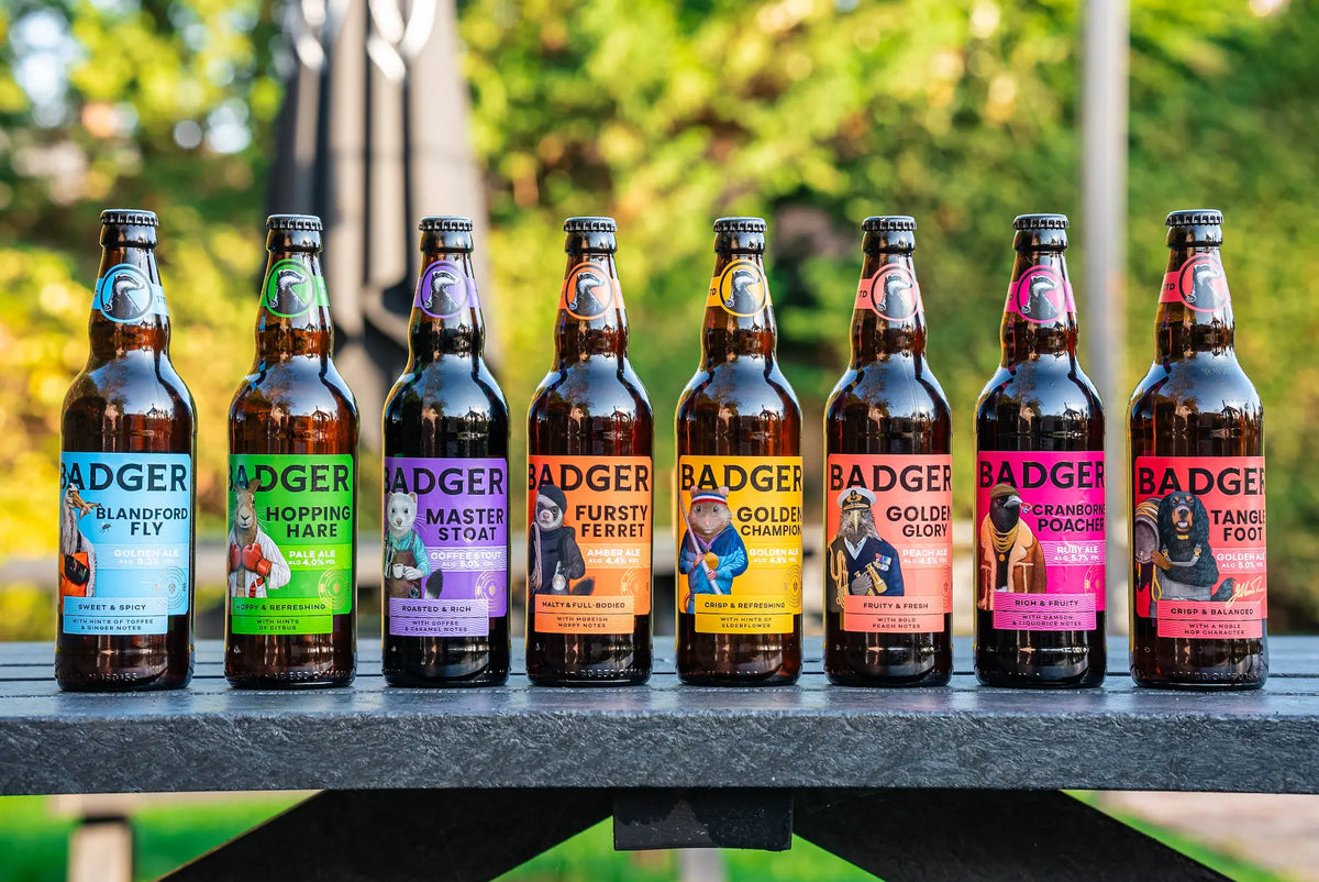 Eight bottles of Badger beer arranged in a row on a dark outdoor table, with a blurred green garden background.