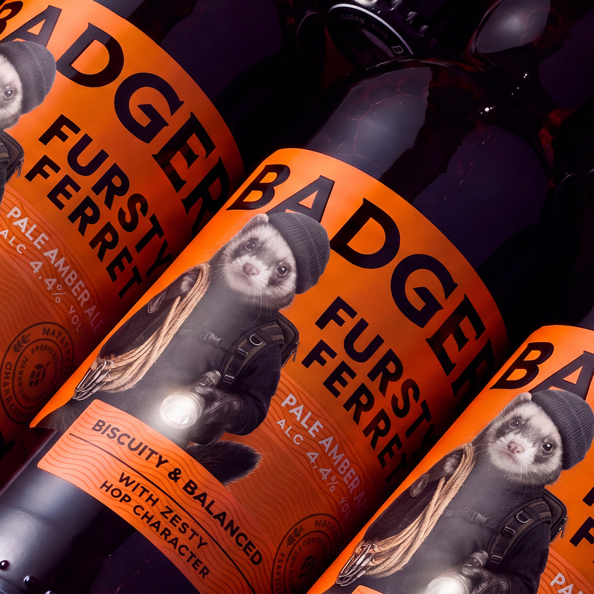 Three dark glass bottles of Badger Fursty Ferret Pale Amber Ale lined up diagonally, featuring bright orange labels with a graphic of a ferret in a beanie and jacket.