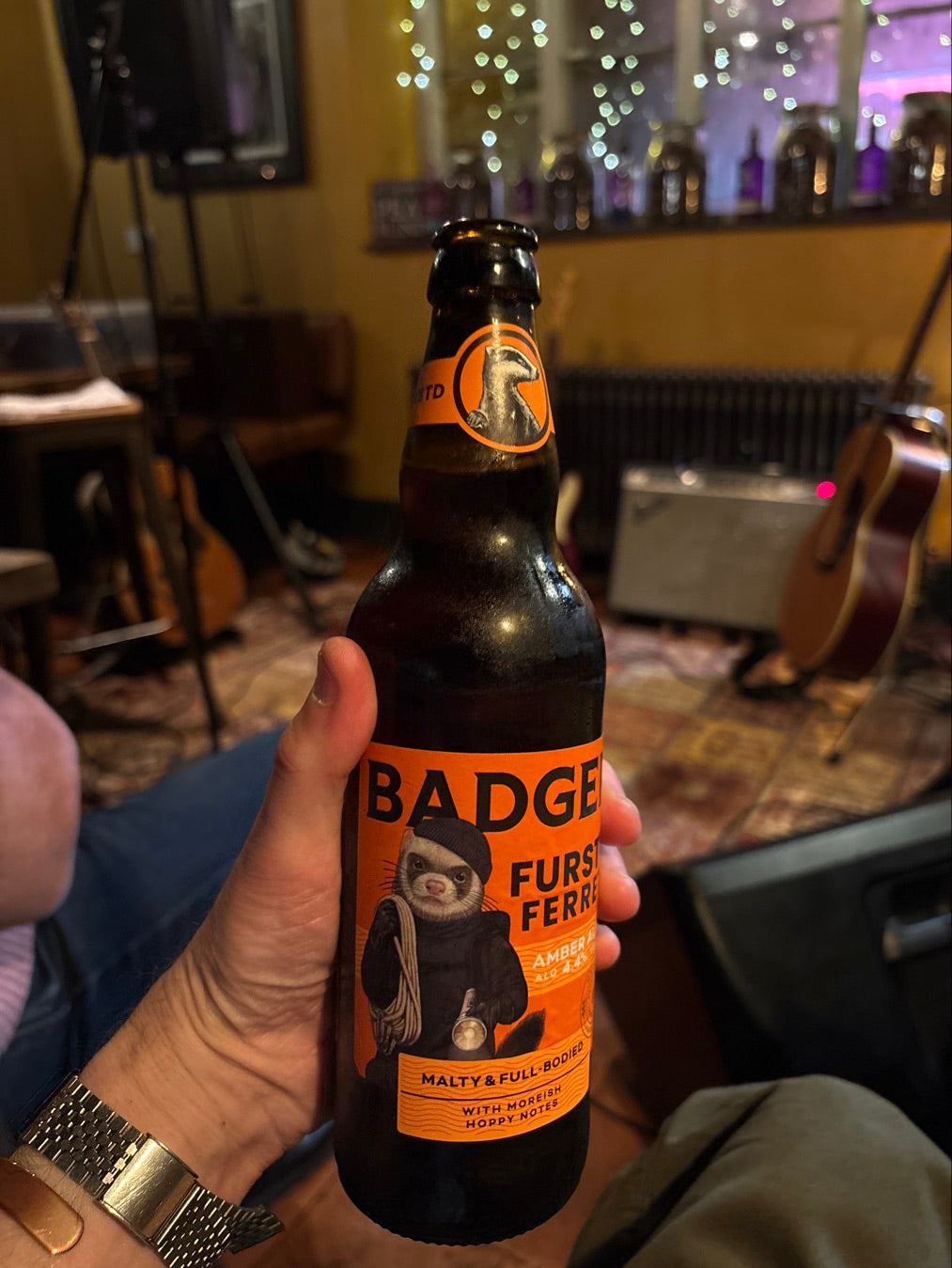 A hand holds a dark brown bottle of Badger Fursty Ferret Amber Ale against a blurred indoor background with guitars and warm lighting. The bottle label is bright orange with black text and an illustration of a ferret figure.