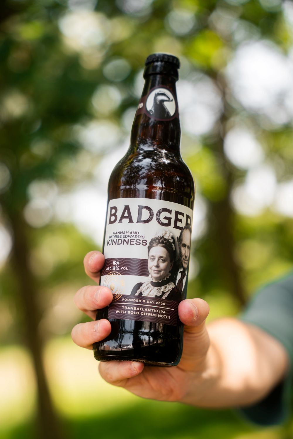 A hand holding a dark brown glass bottle of Badger Hannah and George Edward's Kindness IPA against a blurred outdoor background of green trees.