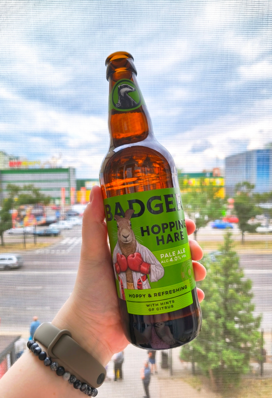 Person holding bottle of Badger Hopping Hare in front of window, with stores, units and cars in the background across the road