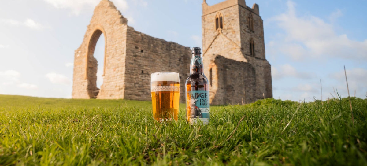 Bottle and pint of Badger Iron Hart on grass, in front of castle ruins, with a bright blue sky and soft clouds as a backdrop