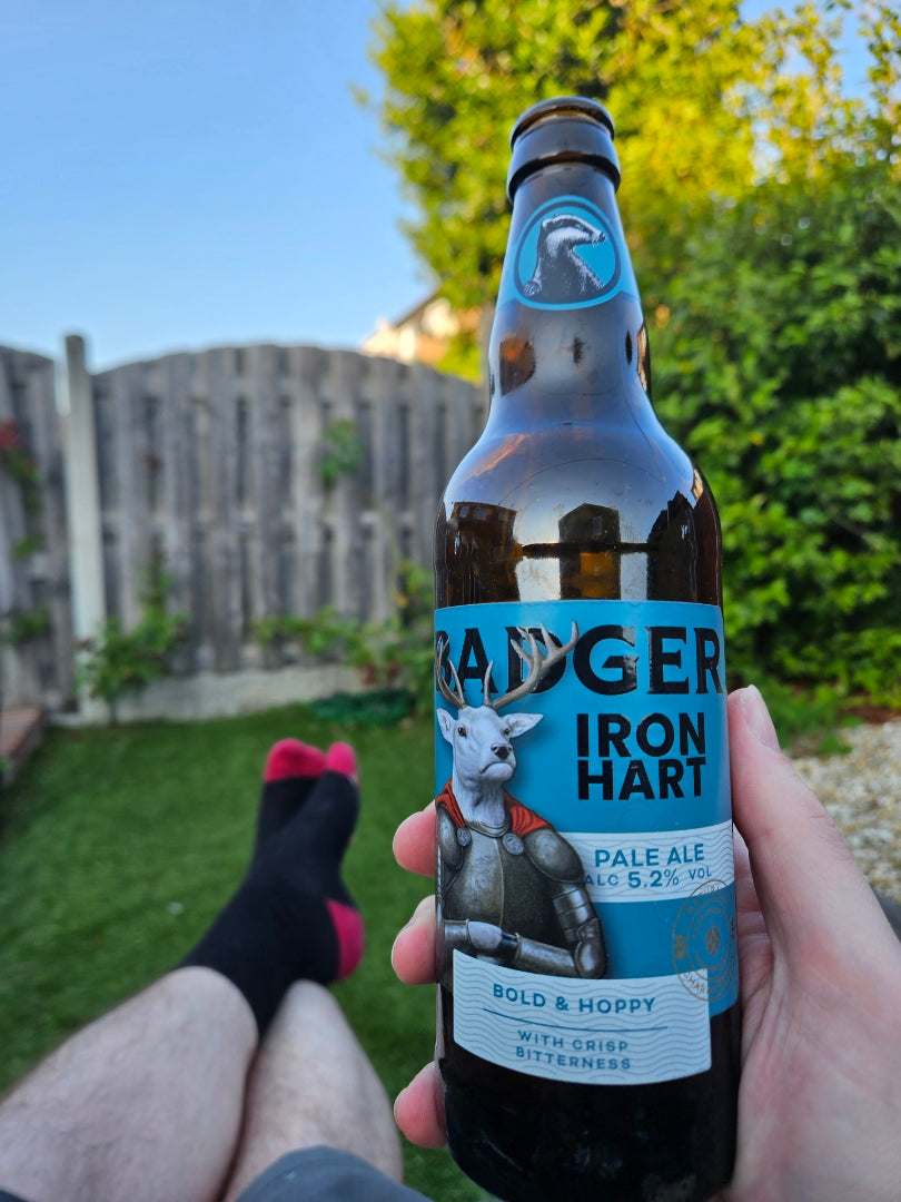 A hand holds a bottle of Badger Iron Hart Pale Ale in a back garden setting, with a wooden fence, green grass, and trees in the background. The person's legs and feet are visible in the lower left, wearing black socks with red toes.