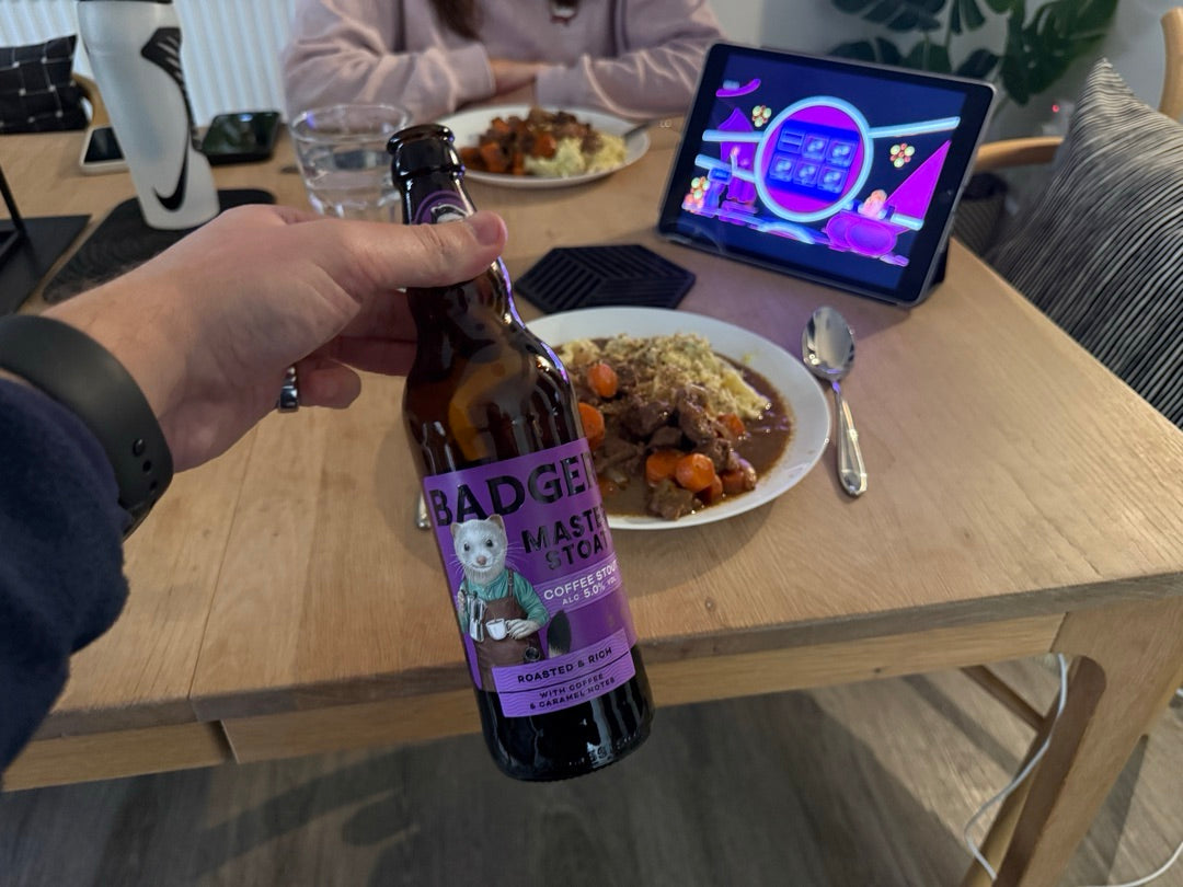 Person holding bottle of Badger Master Stoat in front of plates of food on wooden table, with ipad on table showing sport results