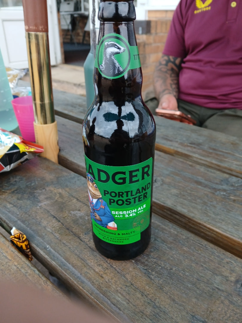 Bottle of Badger Portland Poster on table in pub garden, in front of someone sat on their phone, next to packet of crisps