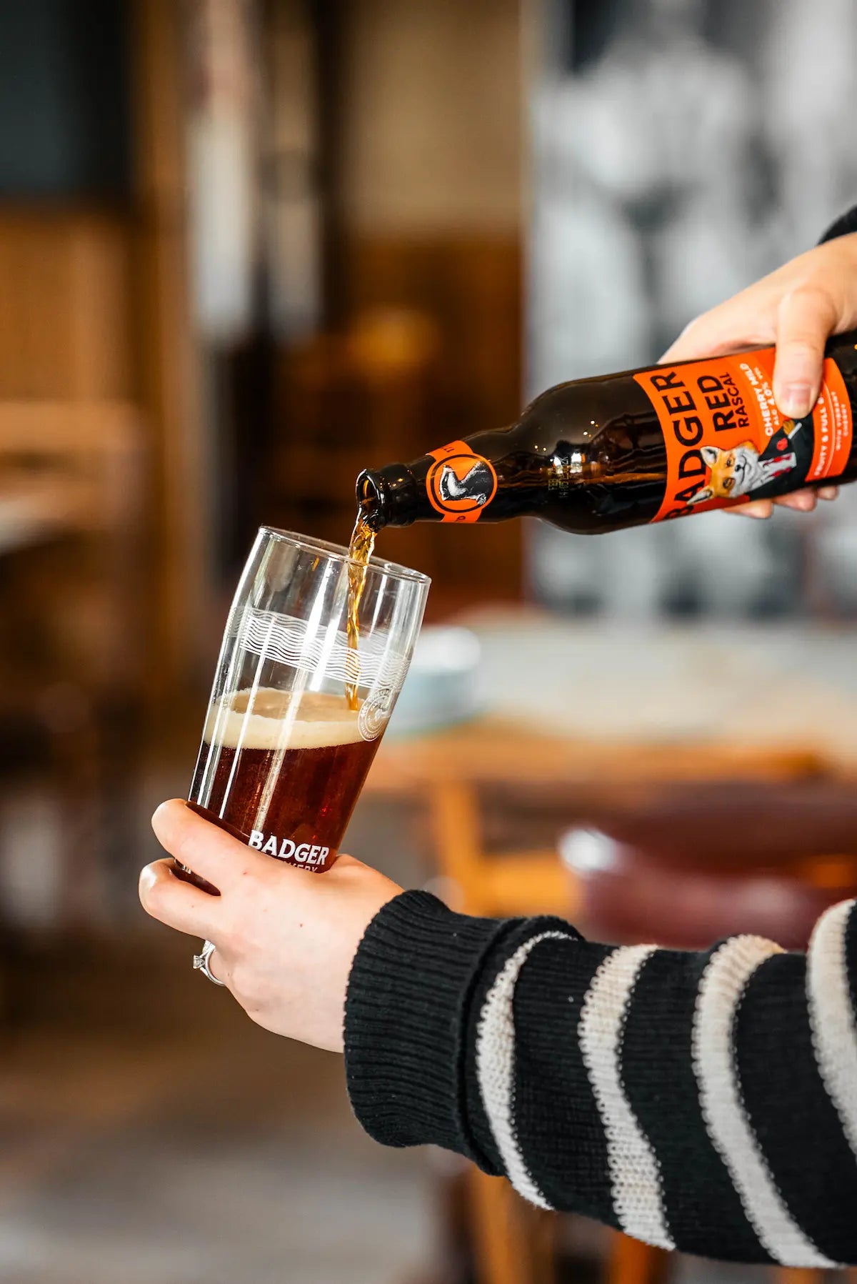 Person pouring bottle of Badger Red Rascal Cherry Mild into a Badger pint glass, with blurred pub room in the background