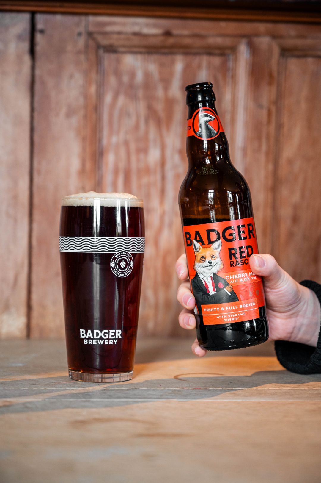 A hand holds a bottle of Badger Red Rascal next to a pint of the dark red beer in a glass with the Badger Brewery logo, set against a rustic wooden background.