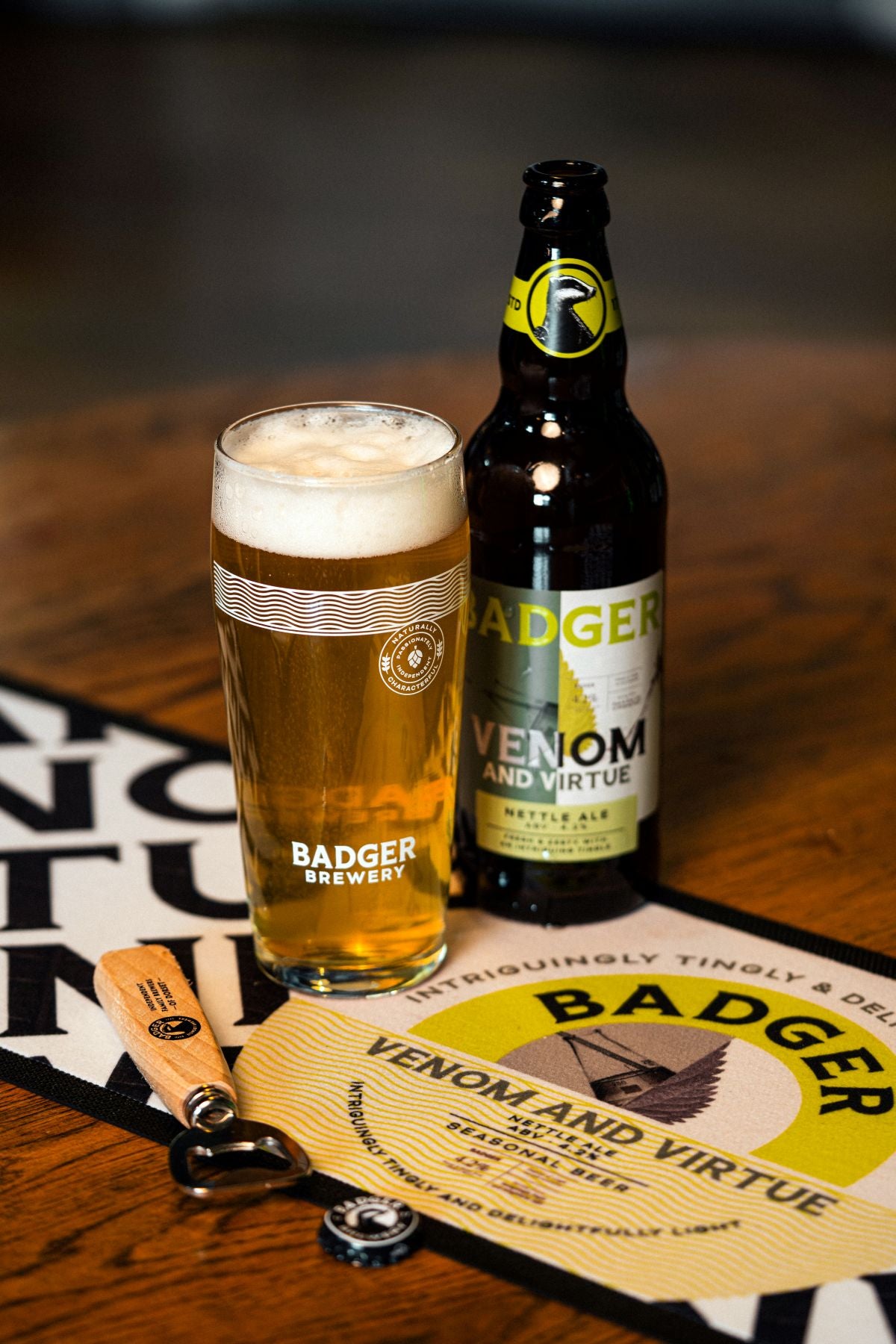 Bottle and pint of Badger Venom and Virtue on a matching bar runner on a wooden table, alongside wooden bottle opener and beer crown.