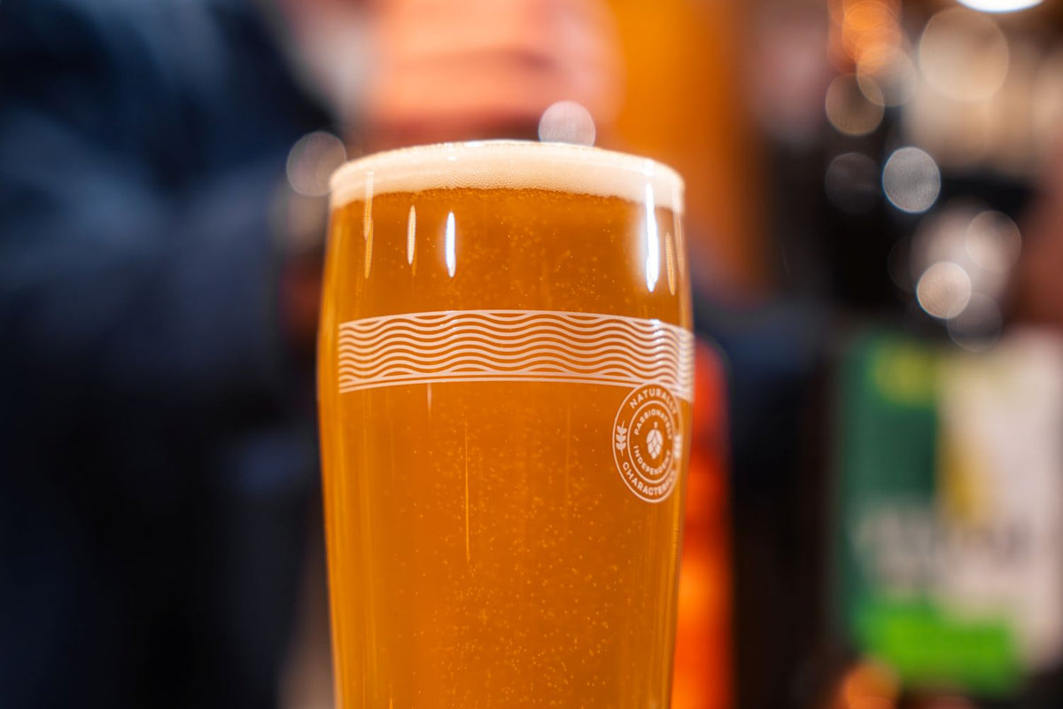A close-up of a pint of Badger Venom and Virtue in a Badger pint glass with a white frothy head, featuring a wave pattern and circular logo on the side, set against a blurred bar background.