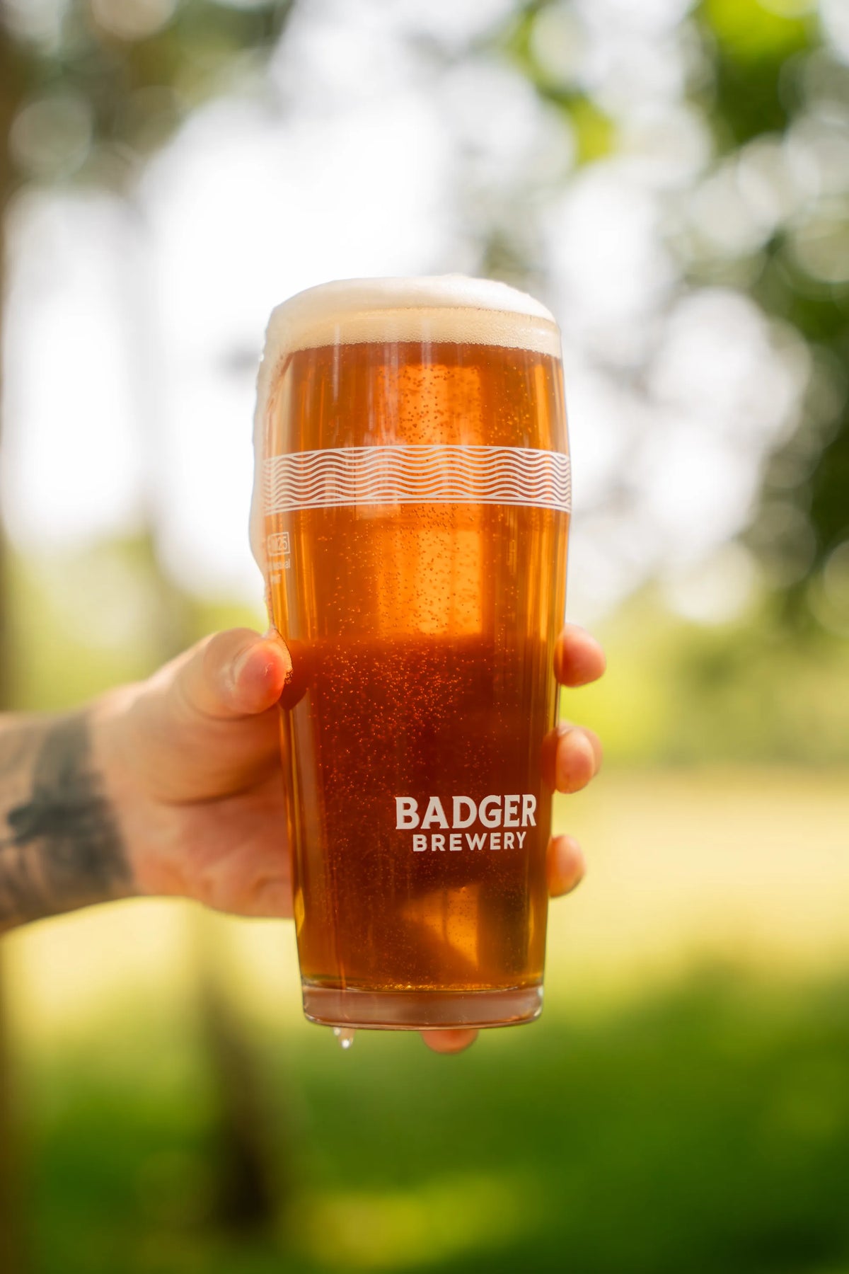 Hand holding a full pint of Badger Kindness IPA outdoors in front of trees