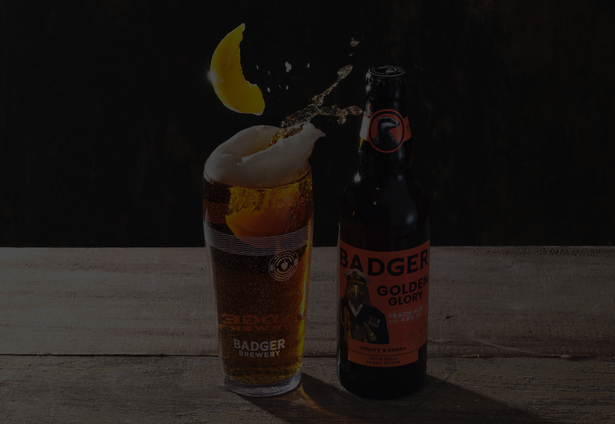 A bottle of Badger Golden Glory stands next to a tall glass of golden ale topped with white foam and a peach wedge falling into the glass, with a splash of liquid in midair against a dark background.