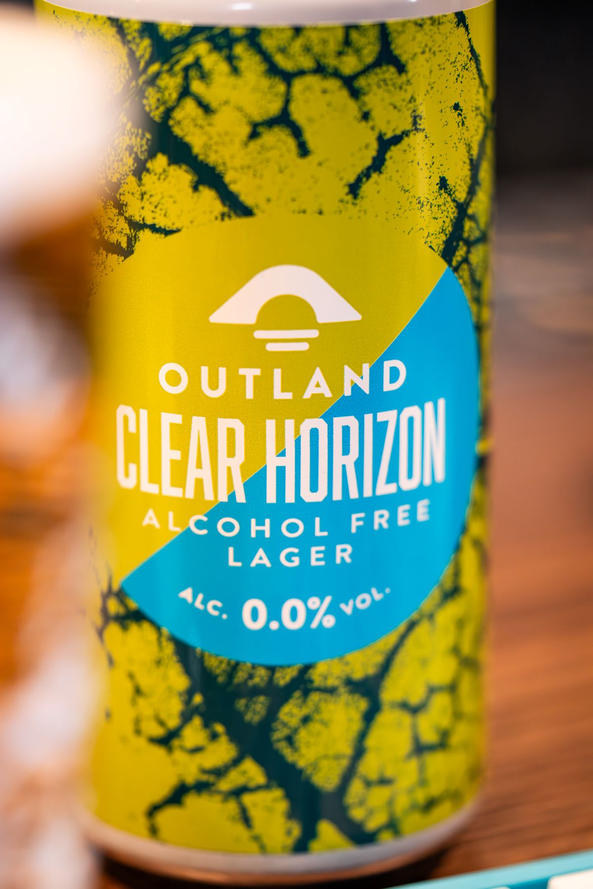 Close-up view of Outland Clear Horizon Alcohol Free Lager - blue and green can, on wooden surface, with blurred edge of pint glass in foreground