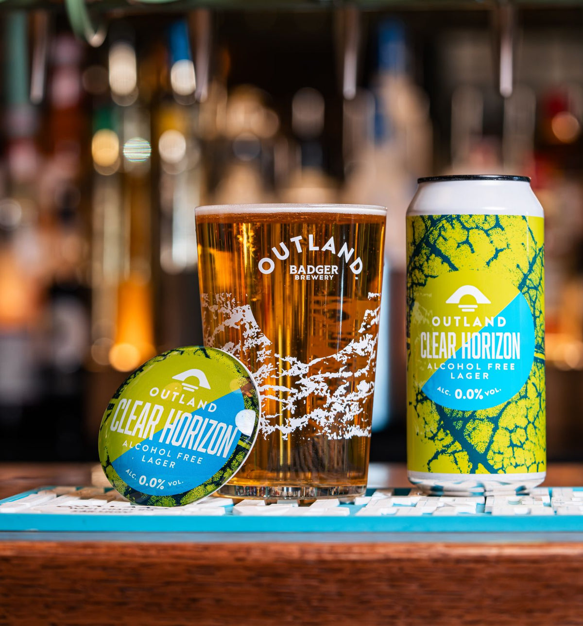 Outland Clear Horizon blue and green fish eye, poured Outland Badger Brewery pint glass and can of Outland Clear Horizon Alcohol Free Lager sitting on blue Outland bar runner, with blurred pub setting in background
