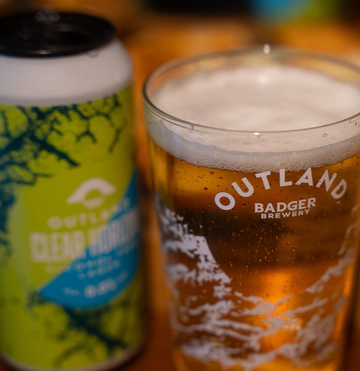 Close-up of Outland Badger Brewery pint glass showing golden liquid, alongside blurred blue and green can of Outland Clear Horizon lager
