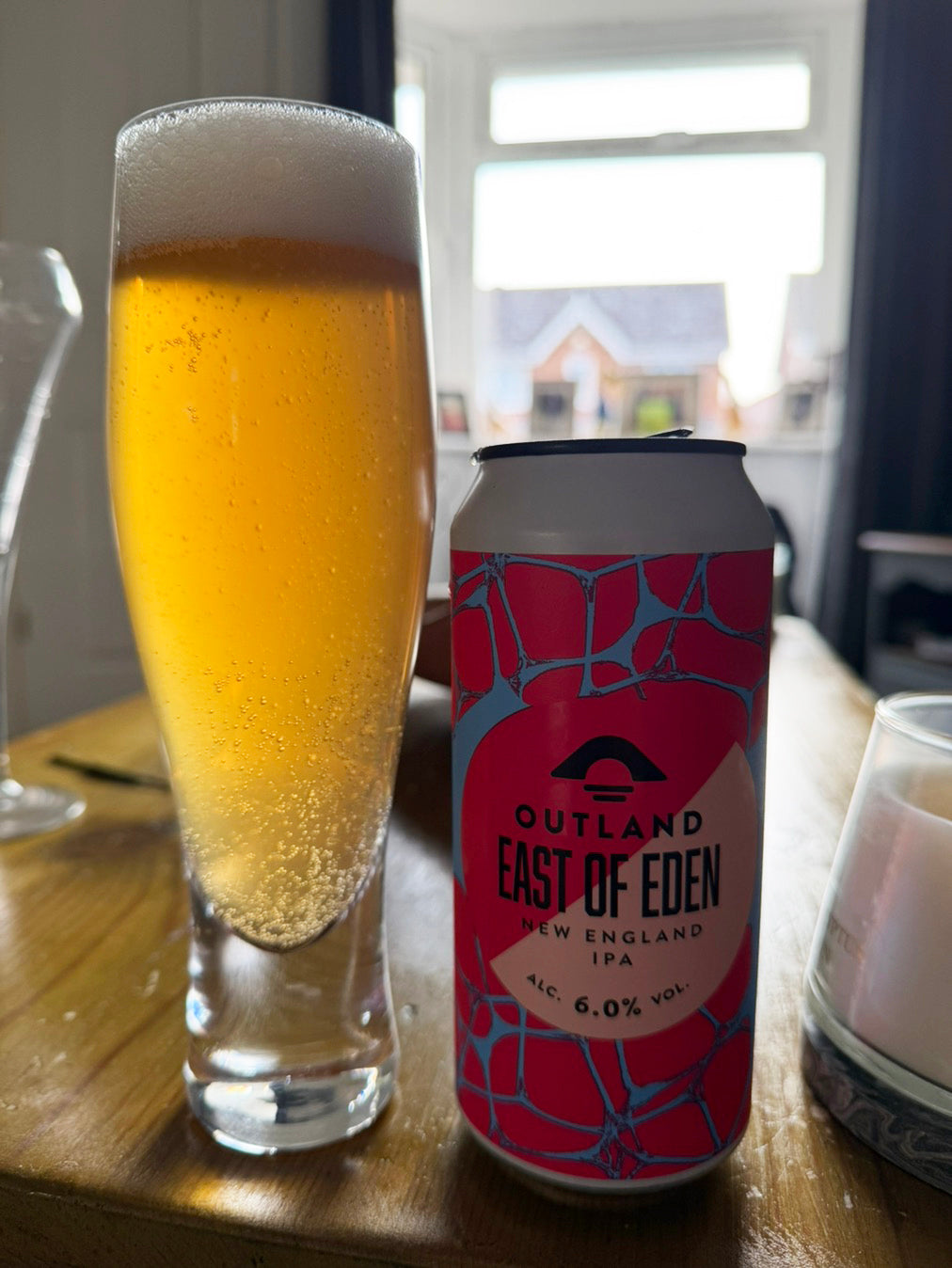 A can of Outland East of Eden New England IPA beer next to a tall glass filled with golden beer and white foam, set on a wooden table in a home interior.