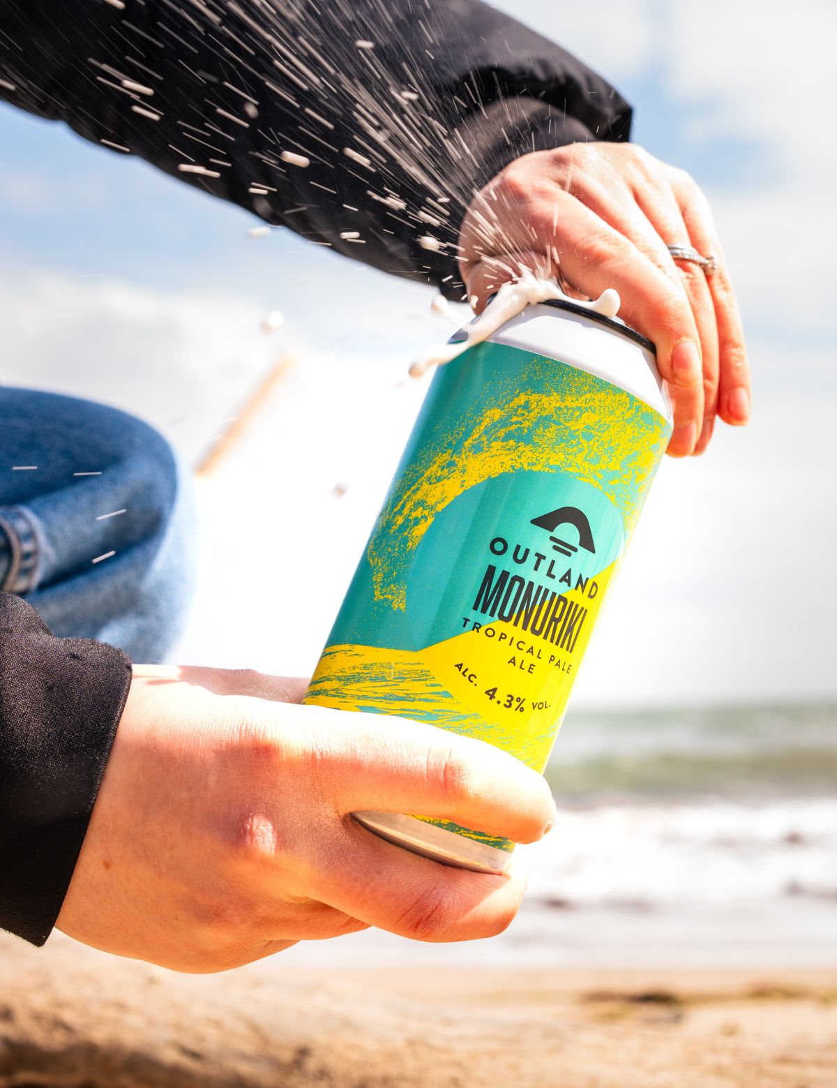 Person opening can of Outland Monuriki teal and yellow can of Tropical Pale Ale craft beer on beach, with dramatic spraying of can