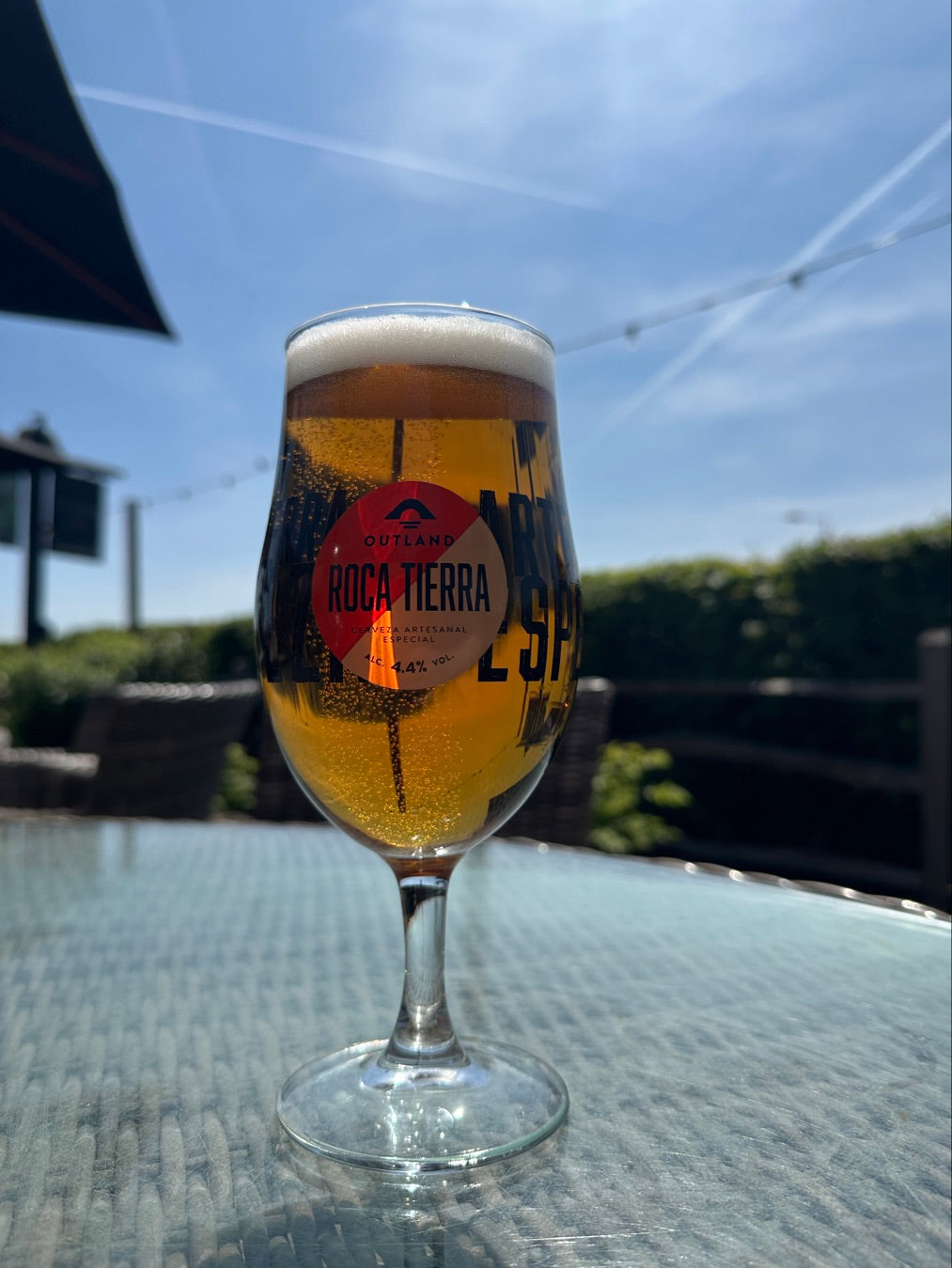 Glass of Outland Roca Tierra on wicker glass topped table in beer garden, with blurred hedges and Hall and Woodhouse pub sign in background