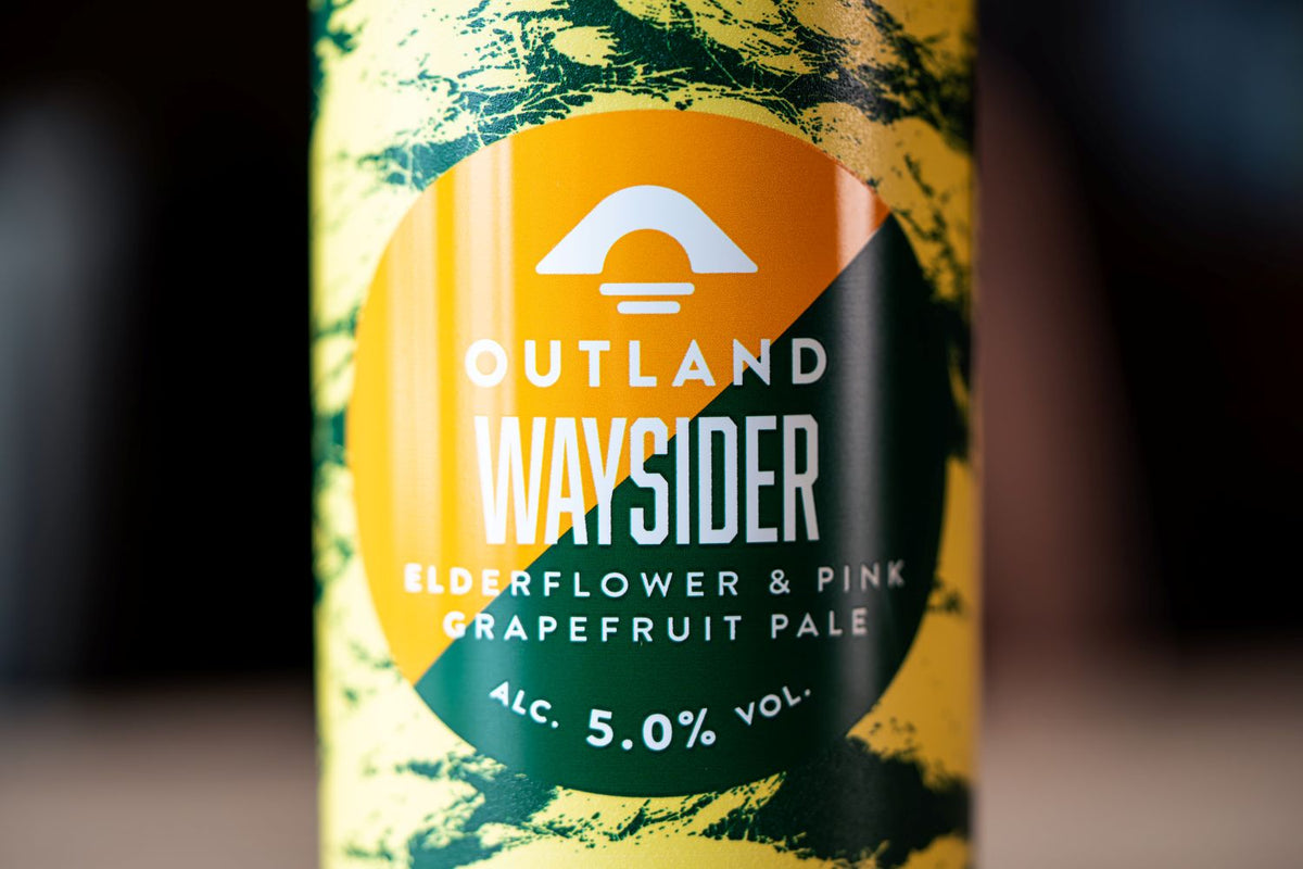 Close-up of can label reading Outland Waysider Elderflower & Pink Grapefruit Pale, with a yellow, orange, and dark green design and blurred dark background.