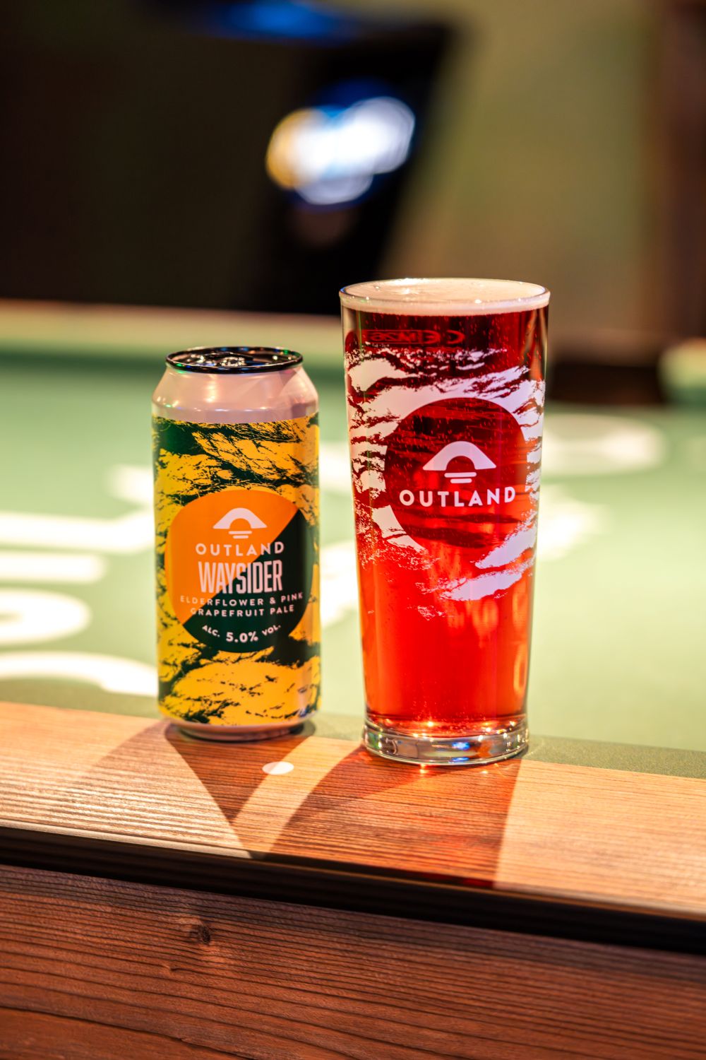 Can and pint glass of Outland Waysider Elderflower and Pink Grapefruit Pale sat on pool table with Outland logo printed on green surface