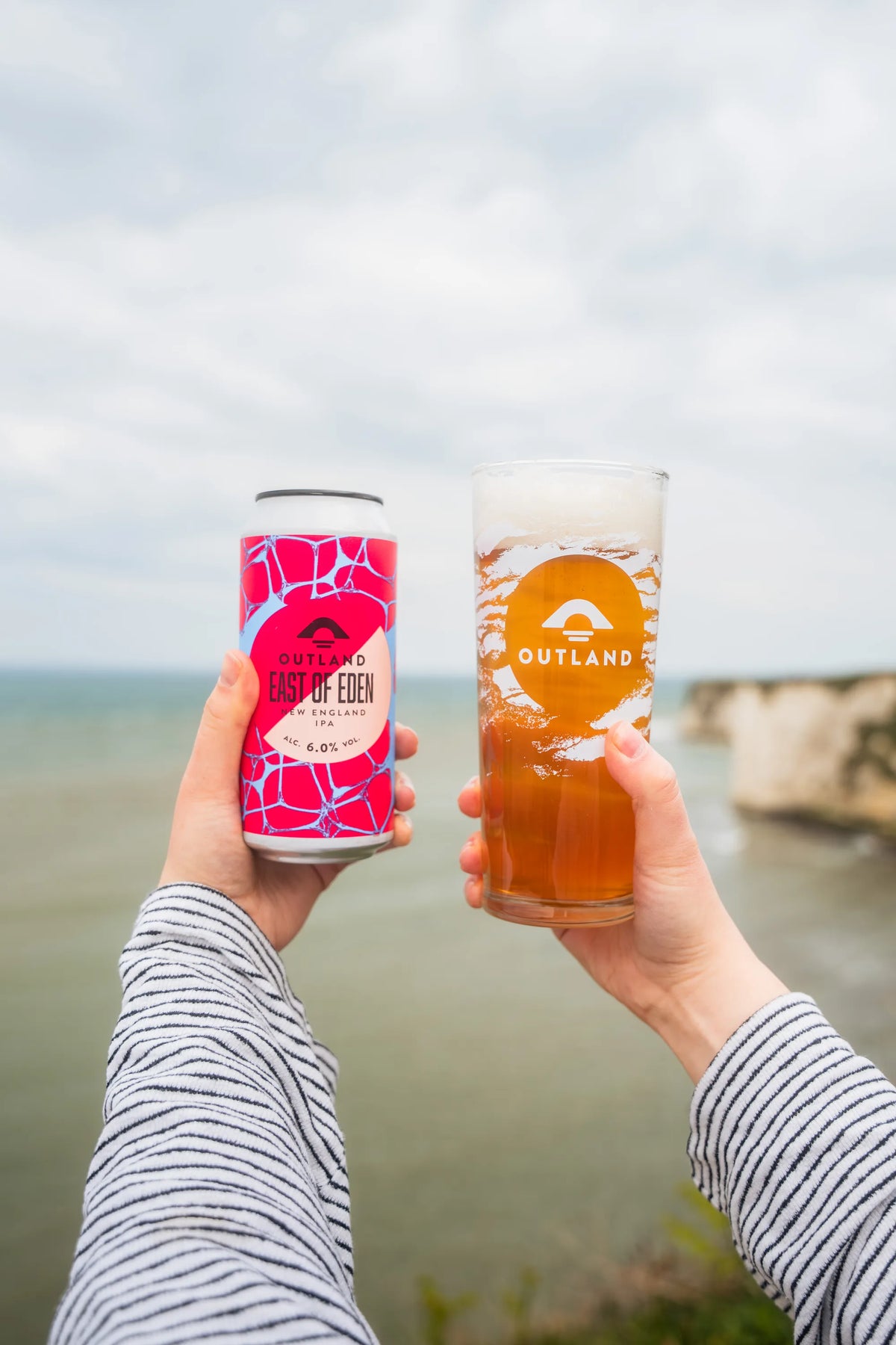 Two hands toasting with cans of Outland East of Eden on the coast path