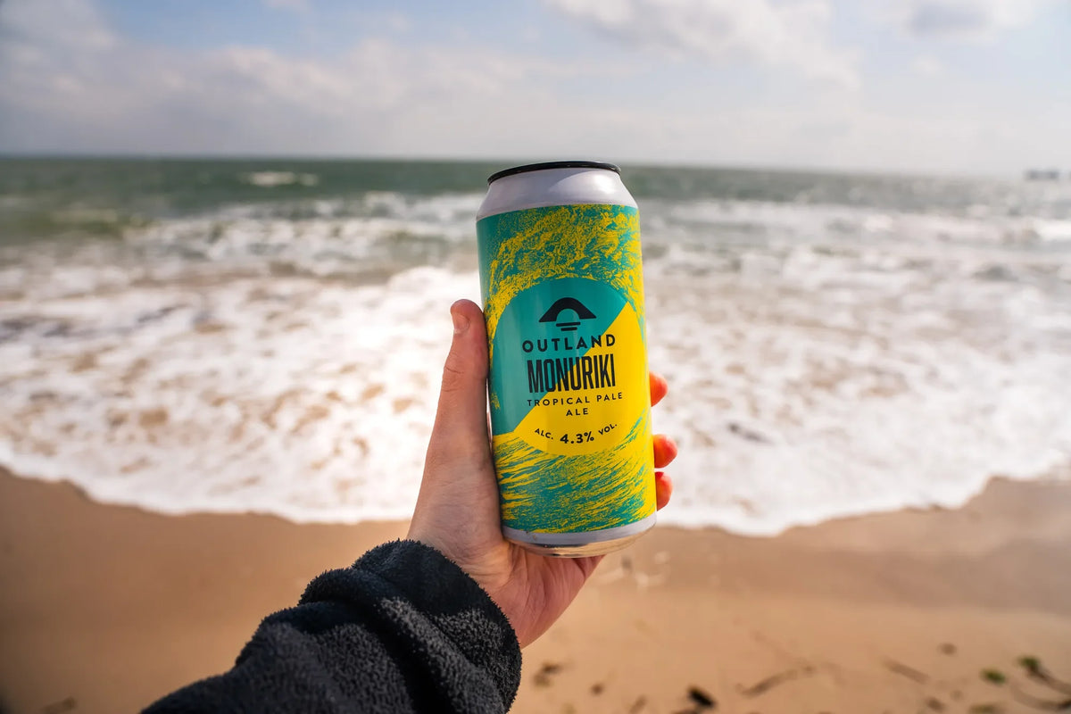 Hand holding a can of Outland Monuriki on the beach with the sea behind