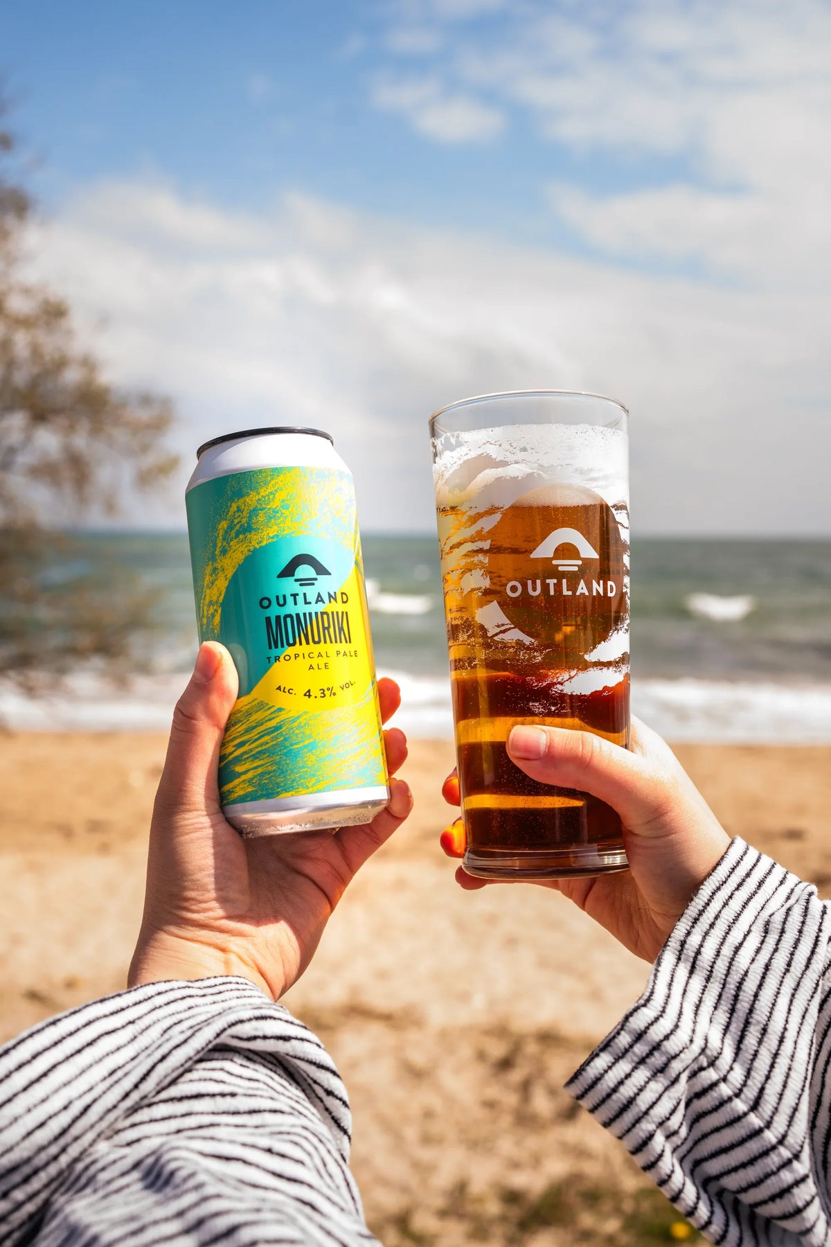 Two hands toasting with a can and a glass of Outland Monuriki on the beach