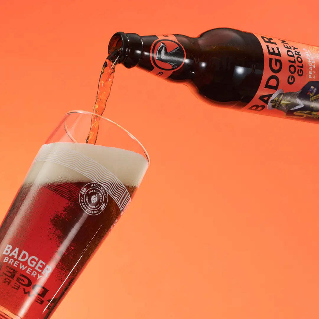 Badger Golden Glory poured from the bottle into a branded glass on a peach background