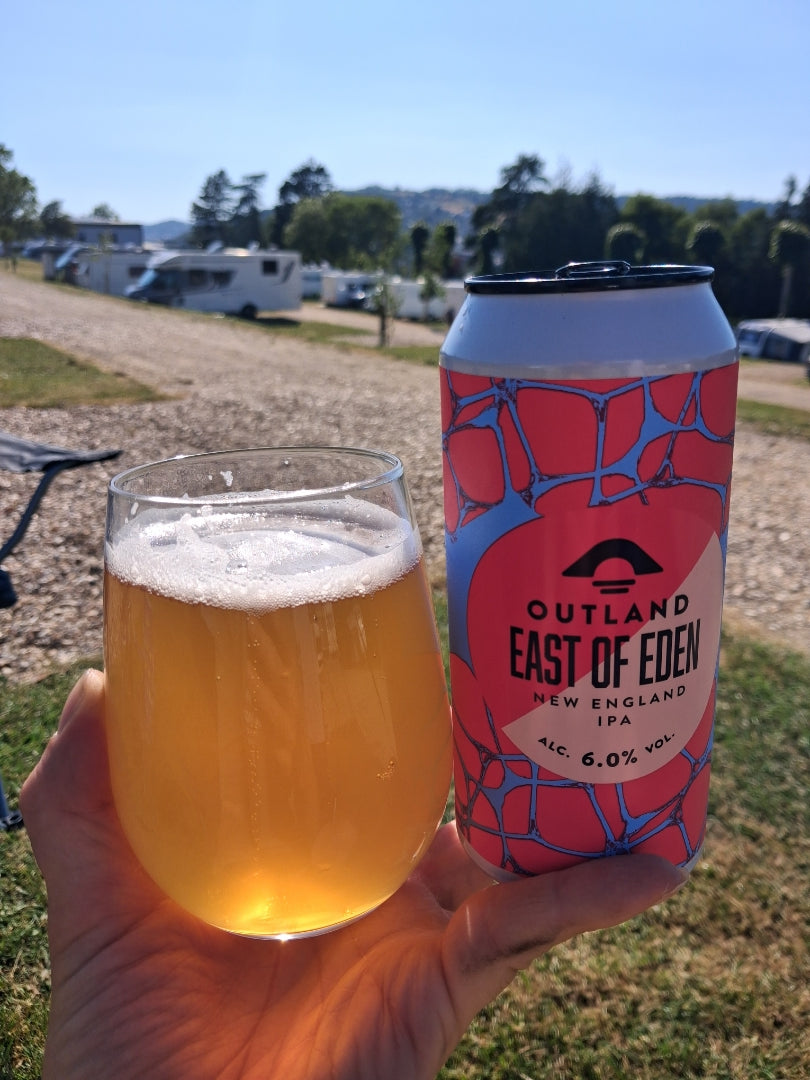 Glass and can of Outland East of Eden held up at a caravan site