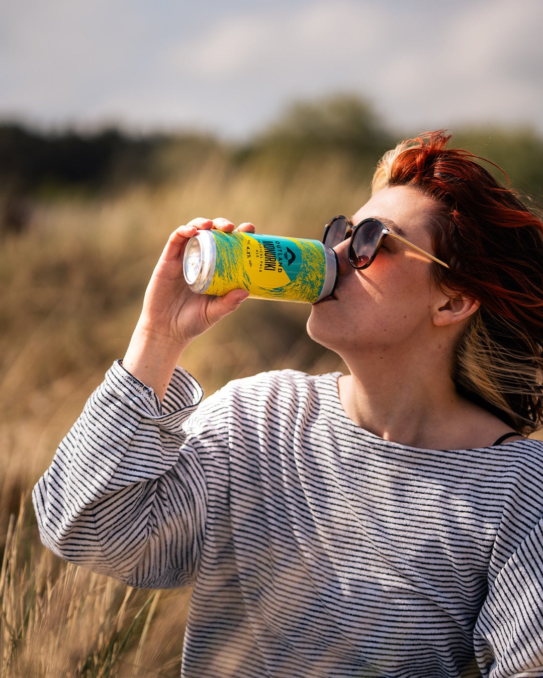 Woman in sunglasses drinking from a can of Outland beer in a field