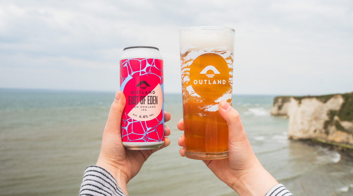 Two hands toasting with a can and glass of Outland East of Eden on the coast