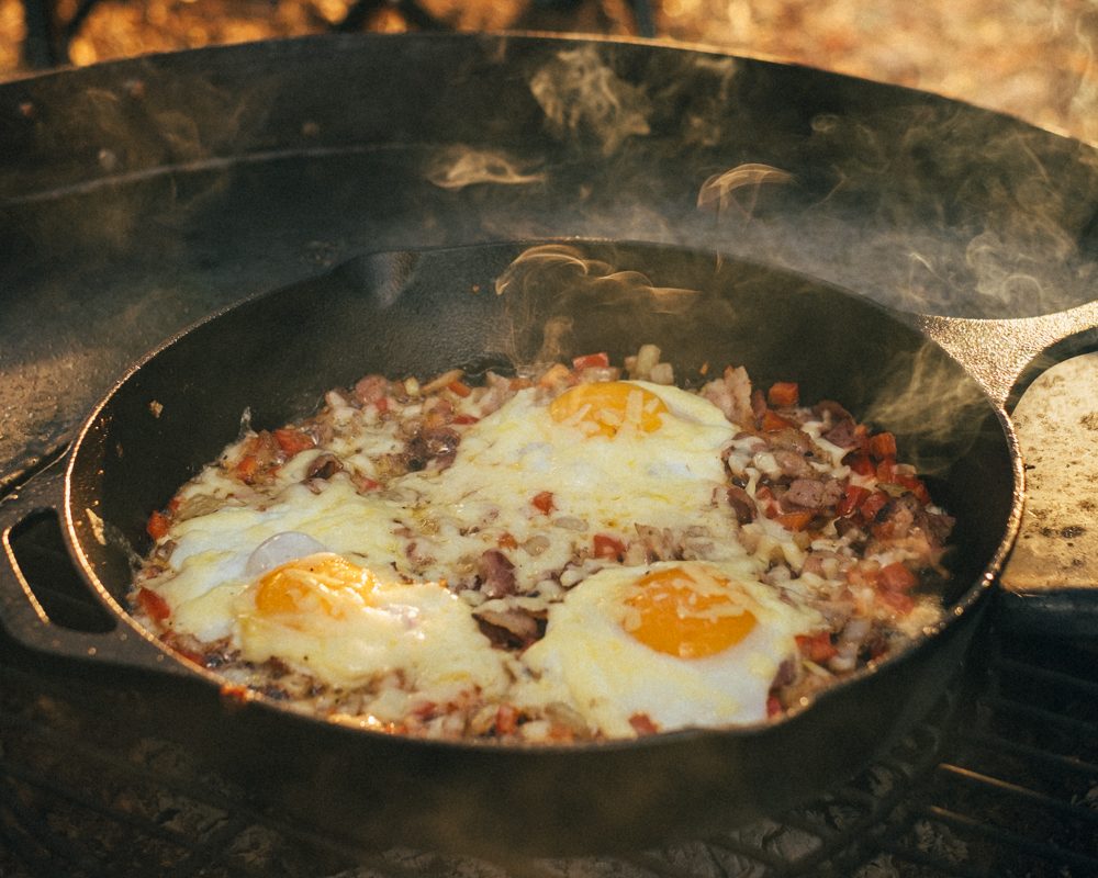 Baked eggs cooking in a cast iron skillet over an open campfire