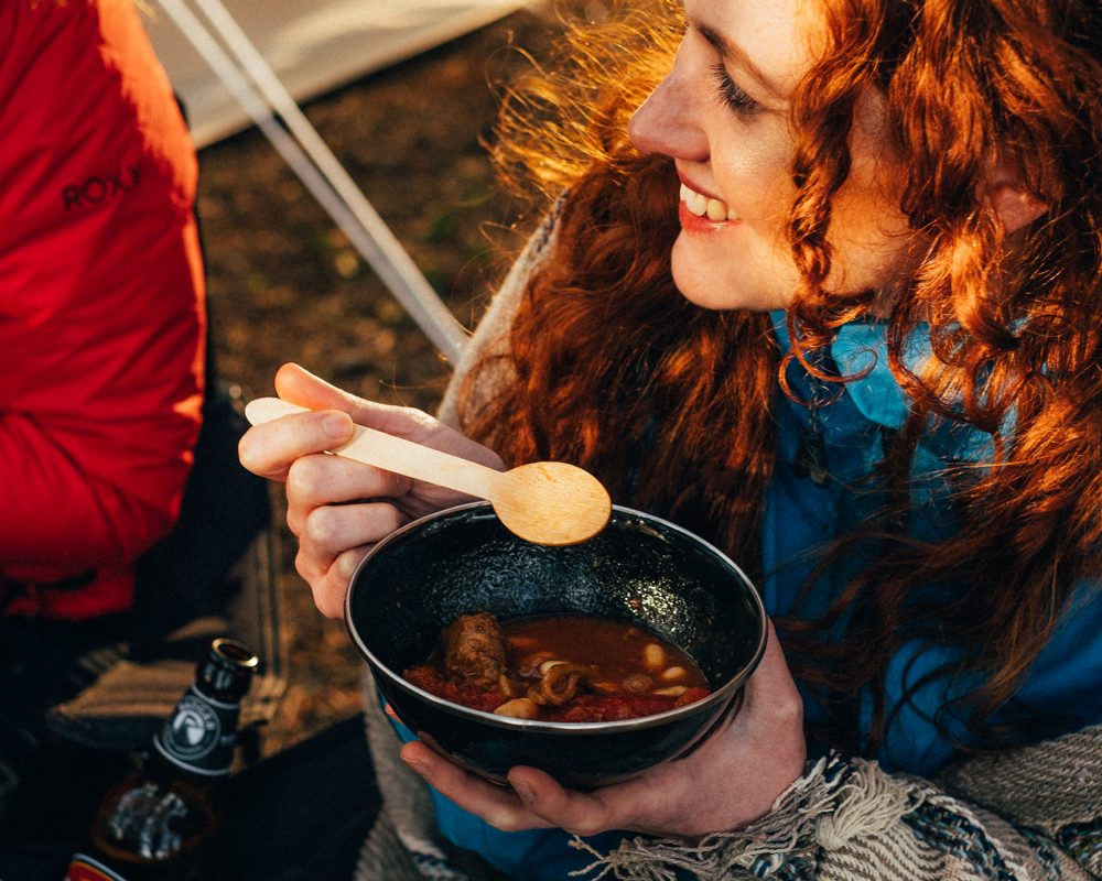 Woman eating campfire hotpot from a bowl outside a tent