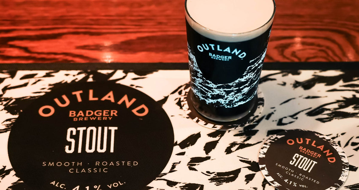 Glass of Outland nitro stout with a creamy head beside branded coasters