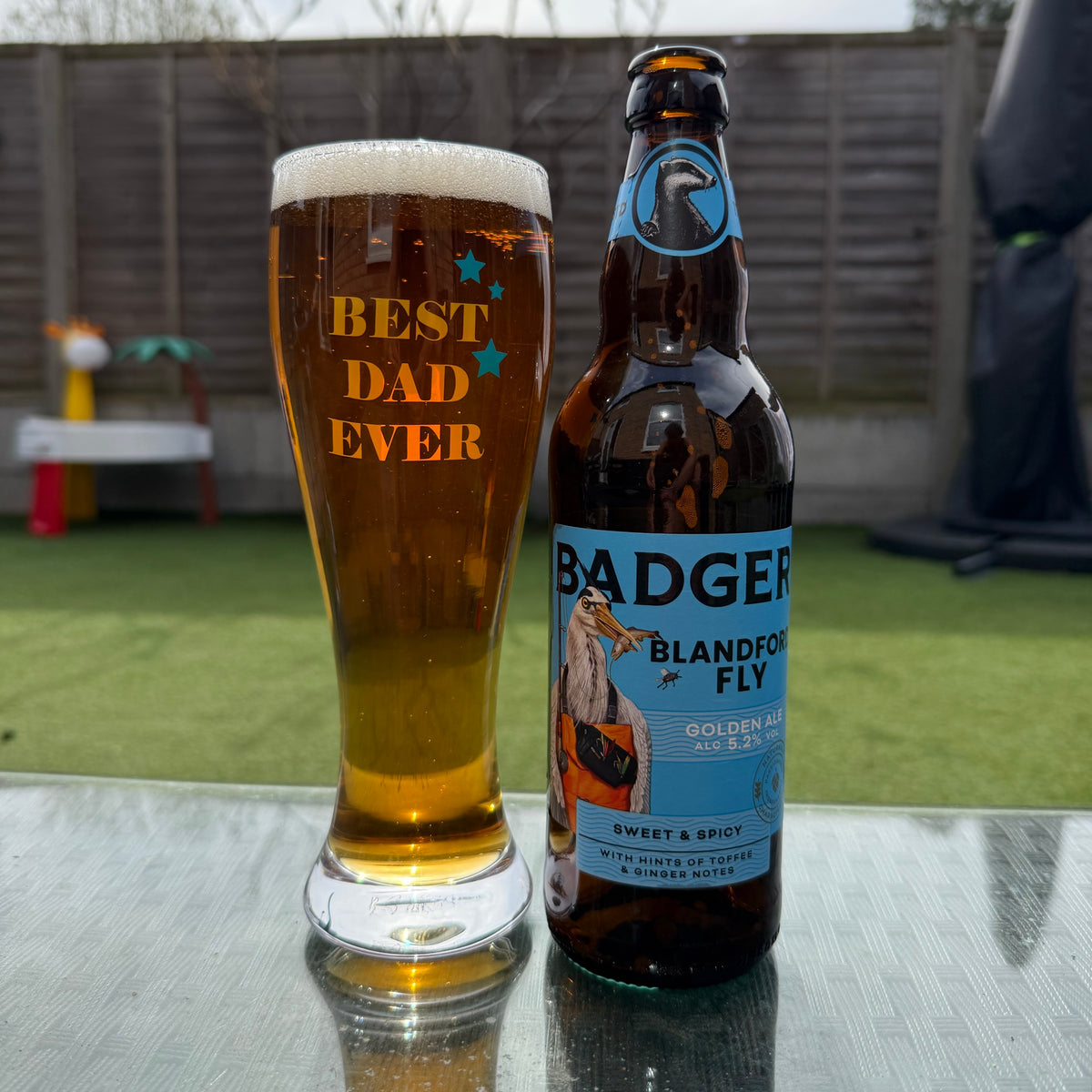 A glass of golden beer with white foam and the words 'BEST DAD EVER' printed on it, standing next to a brown glass bottle labeled Badger Blandford Fly on a reflective outdoor table, in front of blurred garden