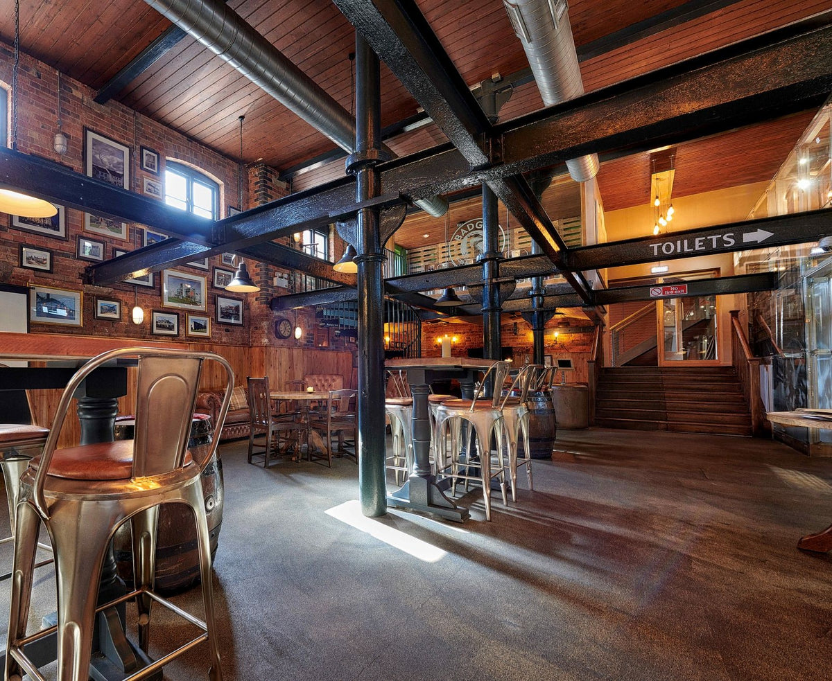 Inside the Badger Brewery tap room in Blandford, Dorset. View of tables and chairs, with industrial style beams along the ceiling and framed portraits on the brick wall. 