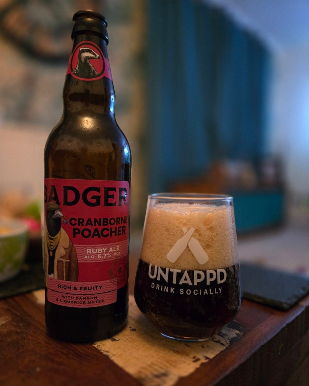 A bottle of Badger Cranborne Poacher Ruby Ale next to an Untappd branded glass of the dark beer with a thick white foam head, set on a wooden table in a dimly lit environment.