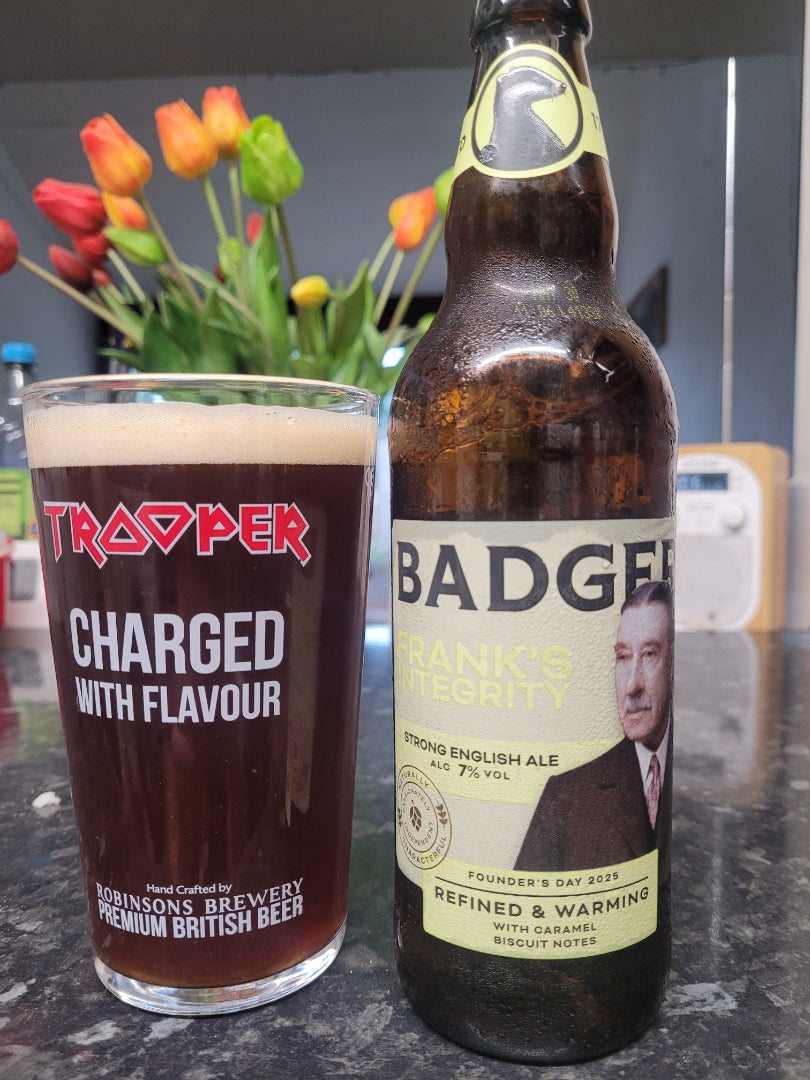A dark brown beer bottle labeled Badger Frank's Integrity stands next to a pint glass filled with dark beer and a thick white head, on a countertop with tulips blurred in the background.