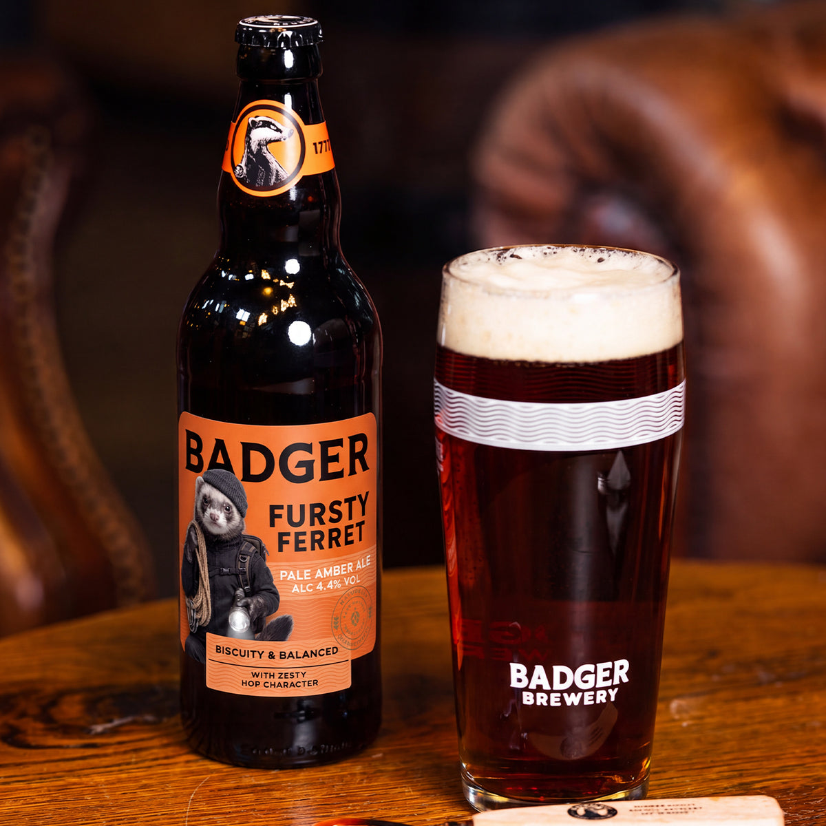 A bottle of Badger Brewery Fursty Ferret pale amber ale next to a glass of the dark, foamy beer on a wooden table, with a blurred brown leather chair in the background.