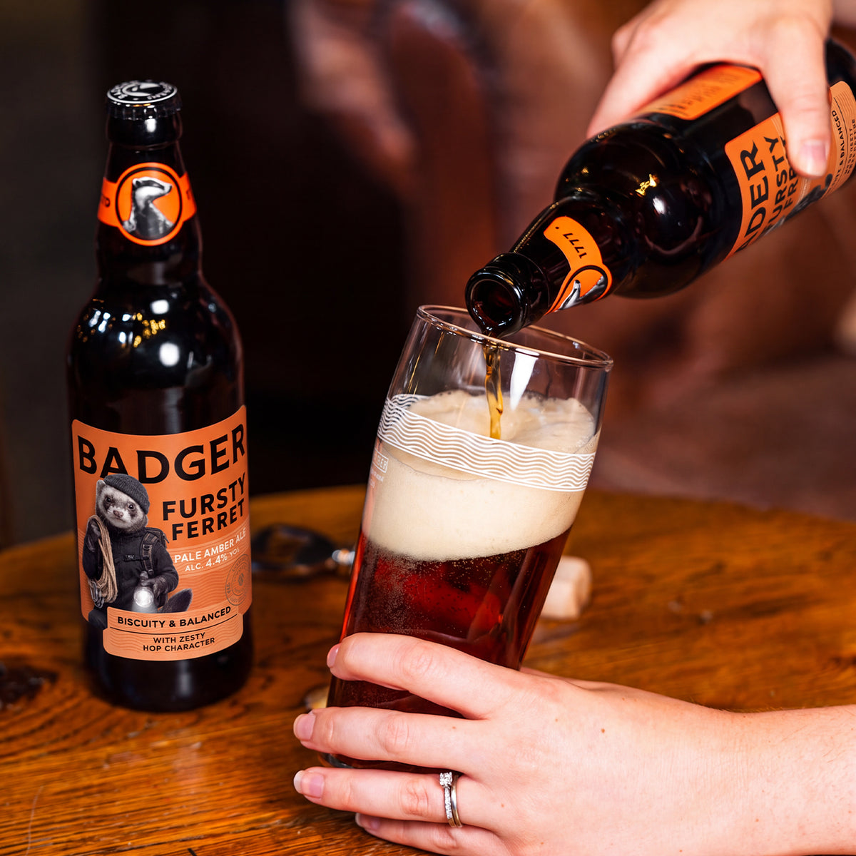 A person pouring a dark amber beer with a thick white head from a Badger Fursty Ferret bottle into a pint glass on a wooden table, with another Badger bottle standing nearby.