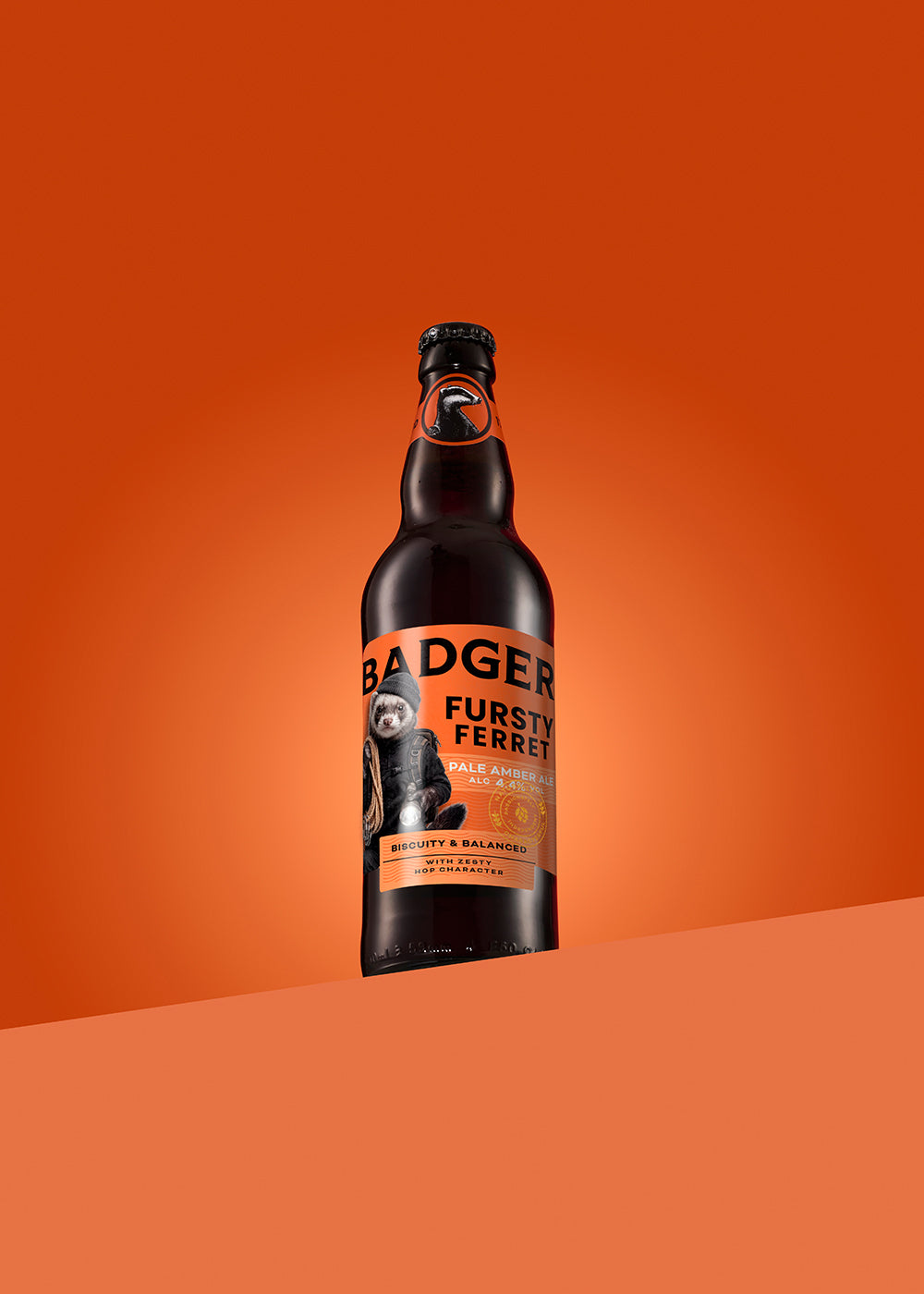 A single bottle of Badger Fursty Ferret Pale Amber Ale stands upright on a curved orange surface against a solid orange background. The bottle is dark amber glass with a black cap and an orange label featuring the brand name and a ferret illustration.