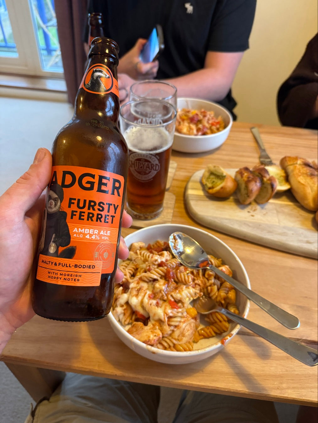 Person holding bottle of Badger Fursty Ferret in front of table with pasta dishes and poured pint glasses, with two other people at table