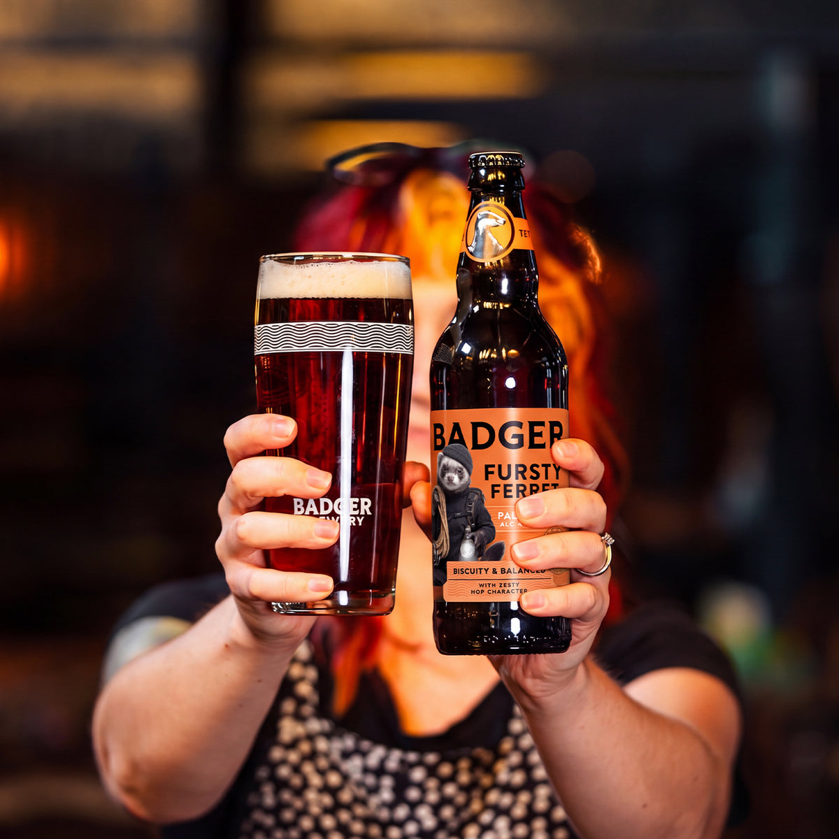 A person holds up a glass of pale amber ale and a bottle of Badger Brewery Fursty Ferret Pale Amber Ale in a dimly lit bar setting.