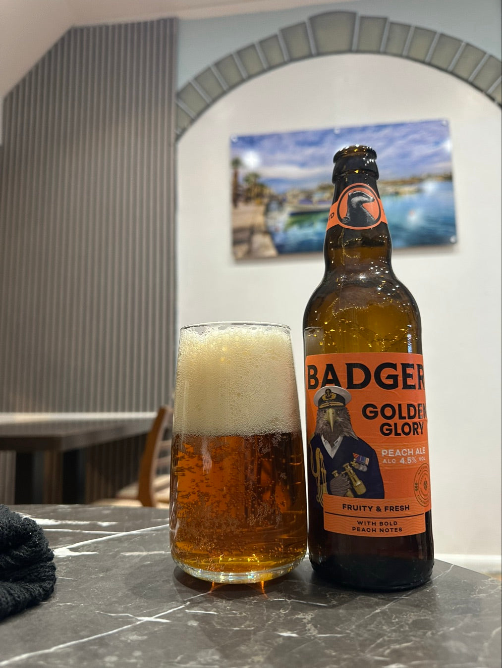 A bottle of Badger Golden Glory beer and a filled pint glass with foam, set on a dark marble tabletop in an indoor setting with a blurred background.
