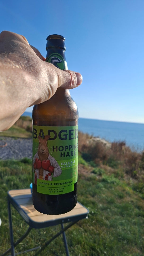 Person holding bottle of Badger Hopping Hare in front of camping table on grass, with views of a blue sky and the sea