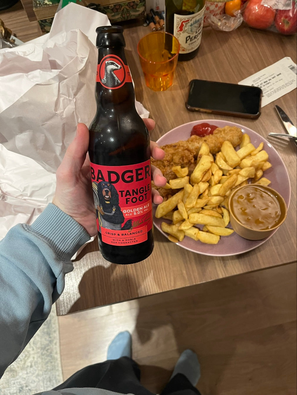 Person holding bottle of Badger Tangle Foot in front of table with takeaway fish and chips on pink plate, paper wrapping and other items on table